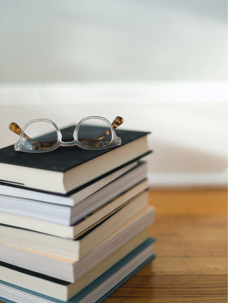 A stack of books on a table with a pair of reading glasses resting on top.